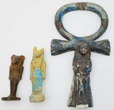 Three Egyptian amulets side by side: two small standing figures in stone and blue faience, and a larger ankh carved with a figure standing within its loop